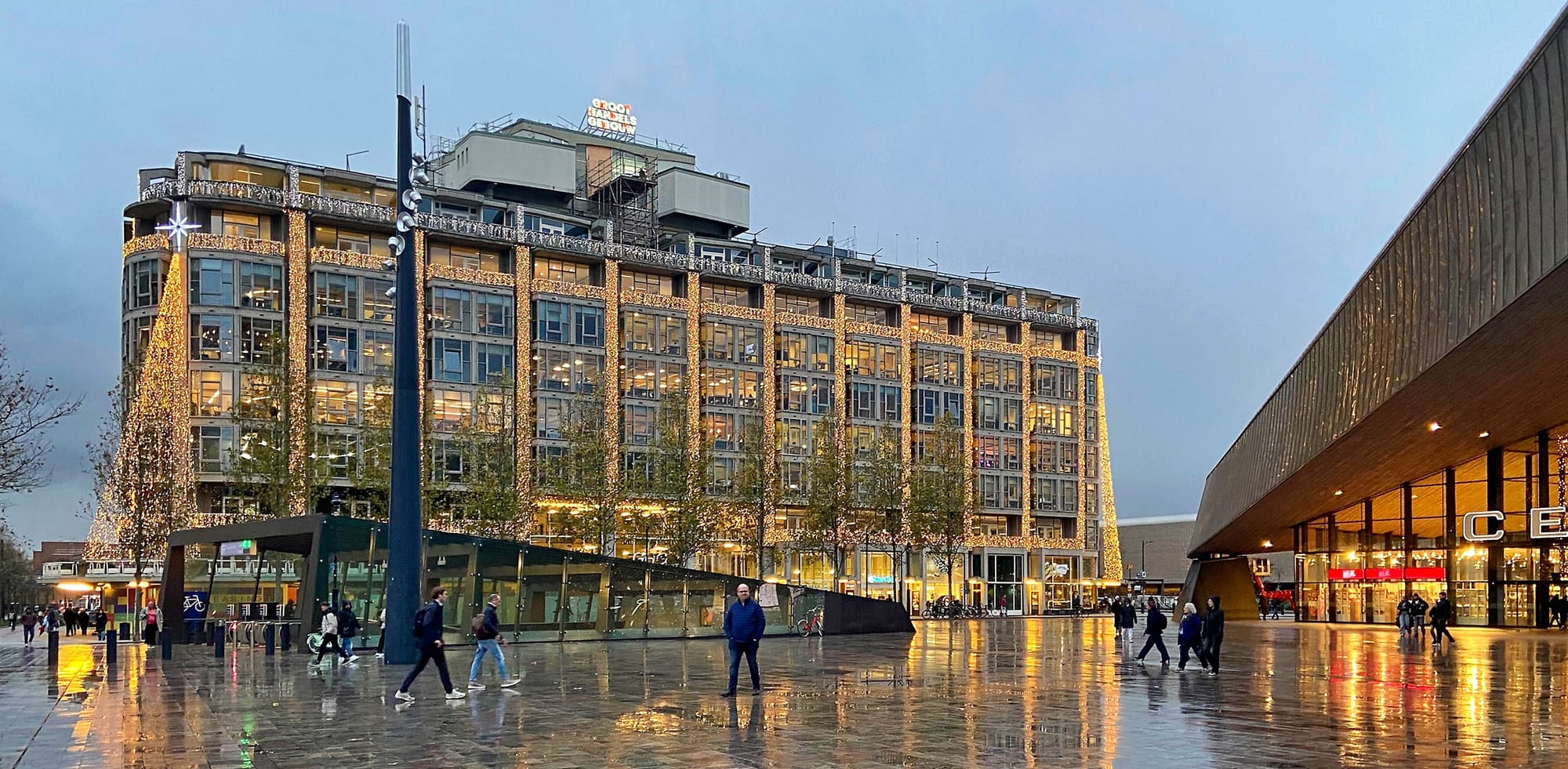 Foto van een plein met een gebouw met allemaal feestverlichting, en een deel van een schuin dak van een station.